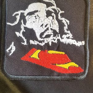 Puede incluir: Parche bordado con fondo negro. El parche presenta un diseño de calavera blanca estilizada sobre un sombrero rojo y amarillo. El parche tiene un borde negro y es probablemente para ropa o accesorios.