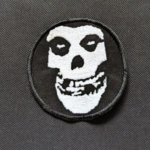 May include: A black and white embroidered patch featuring a skull design. The skull has a wide grin and is set against a black background. The patch is circular with a black border.