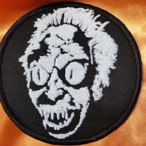 Puede incluir: Parche redondo negro con una imagen bordada en blanco de la cara de un personaje con rasgos exagerados. El personaje tiene ojos grandes, una amplia sonrisa y pelo desordenado. Fondo naranja.