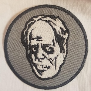 Op de afbeelding: Ronde geborduurde patch met een zombiegezicht in zwart en wit. Het ontwerp heeft een grijze achtergrond en een zwarte rand. Geschikt voor kleding of accessoires.