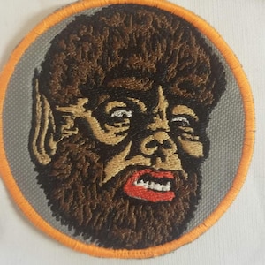 Könnte beinhalten: Gestickter Aufnäher mit einem Werwolfgesicht. Das Design umfasst braunes Fell, beige Gesichtsdetails, rote Lippen und weiße Augen, vor einem grauen Hintergrund. Der Aufnäher ist kreisförmig mit einem orangefarbenen Rand.
