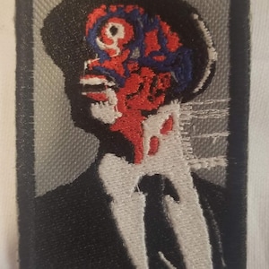 Puede incluir: Parche bordado con una imagen estilizada de una persona con traje y sombrero, con un diseño rojo y azul en la cara. El parche tiene un borde negro y un fondo gris.