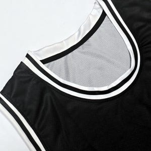 May include: A black sleeveless jersey featuring a round neckline with white and black striped trim. The fabric has a visible mesh texture throughout, and the collar and arm openings are finished with a clean, layered white and black border design.