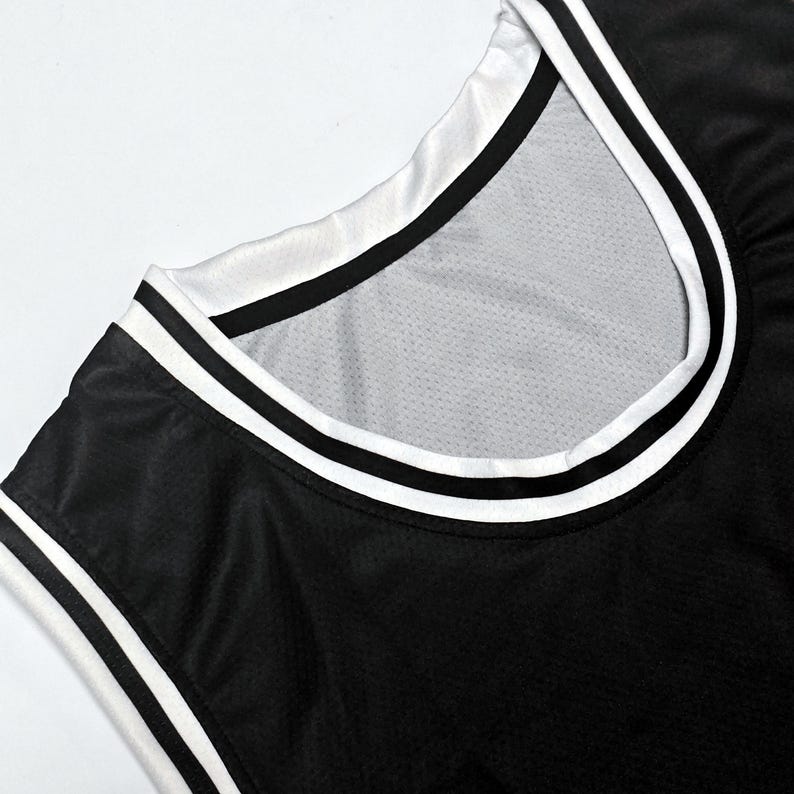 May include: A black sleeveless jersey featuring a round neckline with white and black striped trim. The fabric has a visible mesh texture throughout, and the collar and arm openings are finished with a clean, layered white and black border design.