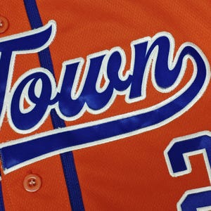 May include: An orange athletic jersey featuring the word TOWN in bold blue script with a white outline. The garment includes a vertical blue stripe, matching orange buttons, and a partial blue number visible on the lower right side of the fabric.