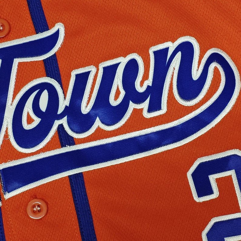 May include: An orange athletic jersey featuring the word TOWN in bold blue script with a white outline. The garment includes a vertical blue stripe, matching orange buttons, and a partial blue number visible on the lower right side of the fabric.