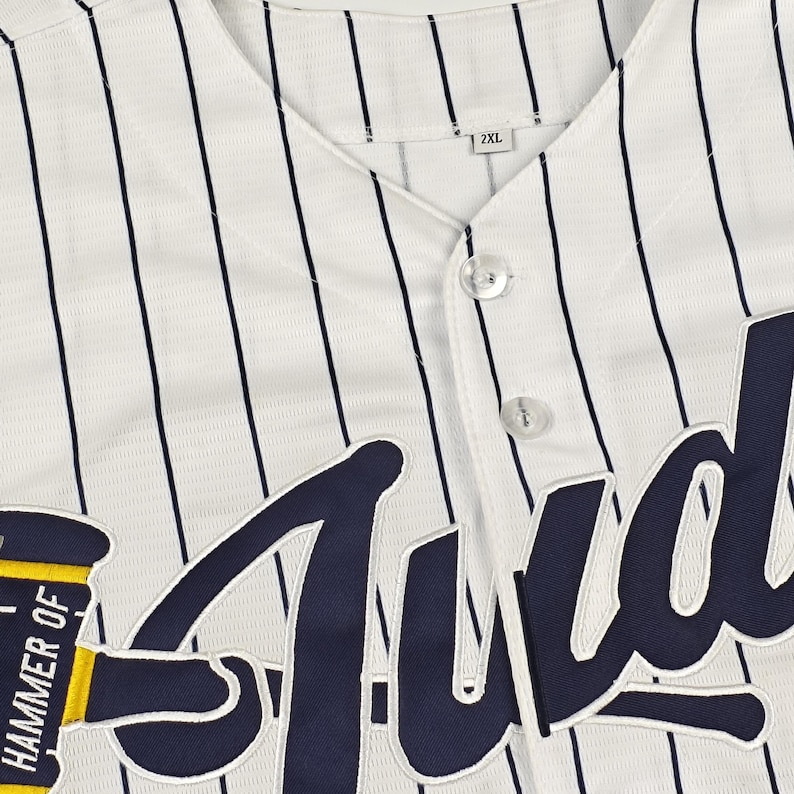May include: A white baseball jersey with thin vertical navy blue pinstripes, featuring a button-down front and a size 2XL tag. The front displays a large navy blue embroidered script logo and a yellow hammer graphic with the text Hammer of.