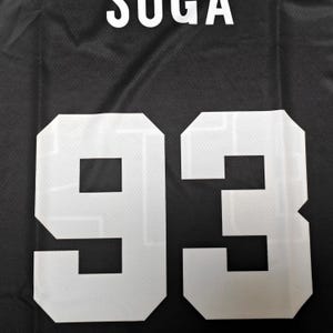 May include: A black athletic jersey featuring the name SUGA printed in white capital letters above the large white number 93. The fabric has a visible mesh texture throughout, and the white graphics are bold and centered on the dark background.