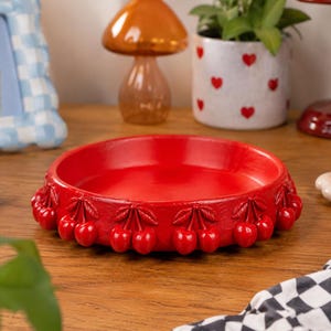 Peut inclure: Un plateau décoratif rond rouge vif avec un motif de cerises sur le bord. Le plateau est fait d'un matériau brillant et repose sur une surface en bois. D'autres objets décoratifs sont en arrière-plan.
