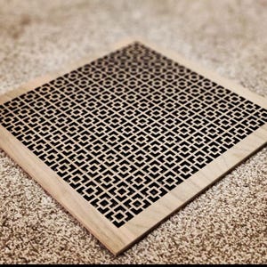 Può includere: Una griglia di ventilazione quadrata in legno con un motivo geometrico tagliato. Il motivo è composto da piccoli quadrati e rettangoli, creando un design a griglia. La griglia di ventilazione è di colore marrone chiaro.