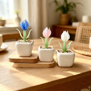 Op de afbeelding: Drie decoratieve tulpen in witte vierkante potten. De tulpen hebben blauwe, roze en witte glazen bloemhoofden met groene stelen en bladeren. De potten staan op een houten dienblad, wat een charmante decoratie creëert.