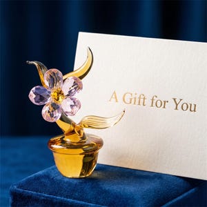 May include: A glass flower figurine with light pink faceted petals and a yellow center, set in a small amber-colored pot. Beside the figurine stands a white card with the gold-colored text A Gift for You printed on the front surface.