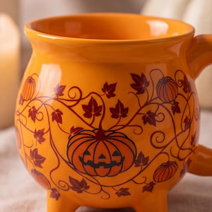 Peut inclure: Un mug en céramique orange de forme ronde rappelant un chaudron, doté de trois petits pieds. L'extérieur présente un motif graphique marron foncé représentant une citrouille d'Halloween entourée de vignes et de feuilles d'automne.