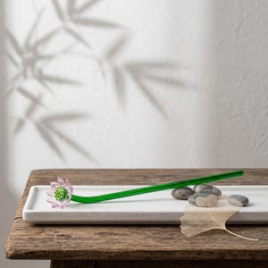 May include: A long, slender green glass stirring rod featuring a delicate, translucent pink lotus flower at one end. The flower has a bright green center. The rod is positioned horizontally on a rectangular white tray with smooth, rounded edges.