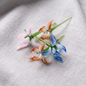 Peut inclure: Trois fleurs en verre fabriquées à la main avec de longues tiges vertes. Une fleur présente des pétales rose clair, une autre des pétales orange et blancs, et la troisième des pétales bleus et blancs. Chaque fleur a une structure centrale en verre vert.