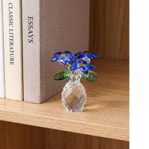May include: A clear crystal figurine of a small vase containing three blue flowers with yellow centers and green leaves. The vase has a faceted, diamond-like texture, and the flowers are crafted from deep blue crystal components with green crystal foliage.