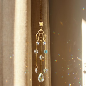 Op de afbeelding: Een goudkleurige metalen zonnevanger met een zonmotief, maansikkels en kleine sterren. Aan de structuur hangen drie fijne gouden kettinkjes, versierd met iriserende, gefacetteerde glaskristallen in de vorm van sterren, rondjes en een druppel.