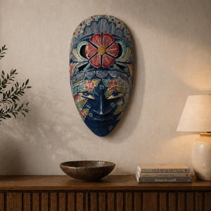 May include: A blue, floral-patterned mask is displayed on a wall. The mask has a central red flower. A vase with greenery, a wooden bowl, a lamp, and books sit on a wooden cabinet. The room is illuminated by the lamp.