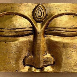 May include: A rectangular, gold-coloured sculpture of a serene face with closed eyes and a prominent nose. The sculpture features detailed lines and a teardrop-shaped symbol above the eyes, suggesting a spiritual or religious theme.