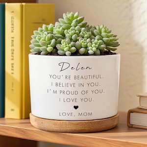 May include: A white ceramic planter with a succulent plant. The planter has the name "Delen" and the message "YOU'RE BEAUTIFUL. I BELIEVE IN YOU. I'M PROUD OF YOU. I LOVE YOU. LOVE, MOM" in black script. It sits on a wooden saucer.