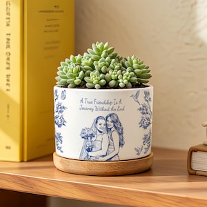 May include: A white ceramic cylindrical planter with a wooden base containing a green succulent. The planter features blue illustrations of two people and floral patterns with the text, A True Friendship Is A Journey Without An End.