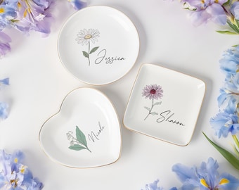 May include: Three white ceramic trinket dishes with gold rims. One round dish features a daisy and the name "Jessica". A square dish has a purple flower and the name "Sharon". A heart-shaped dish displays a lily of the valley and the name "Nicole".