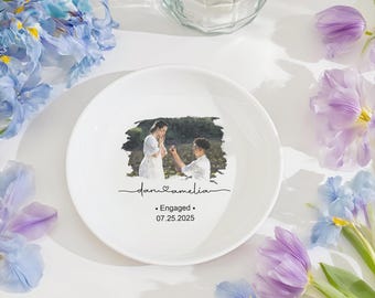 以下が含まれることがあります： カップルの写真がプリントされた白いセラミックプレート。テキストは「dan & amelia」と「Engaged 07.25.2025」です。プレートは紫と青の花に囲まれ、背景にはガラスの花瓶があります。