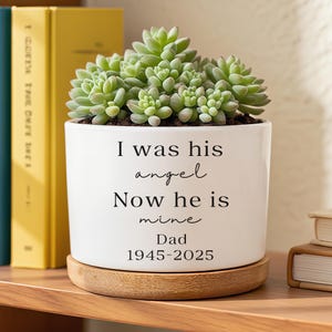 Peut inclure: Un pot de fleurs en céramique blanche avec une plante succulente. Le pot porte l'inscription "I was his angel Now he is mine Dad 1945-2025". Le pot repose sur un dessous de plat rond en bois.