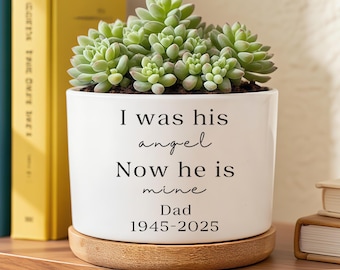 May include: A white ceramic planter with a succulent plant. The planter has the text "I was his angel Now he is mine Dad 1945-2025". The planter sits on a round wooden coaster.