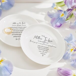 Op de afbeelding: Twee witte keramische ringschalen met zwarte tekst. E&eacute;n leest "Mother of the Bride" met een datum. De andere zegt "Mother In Law" met een bericht en datum. Een gouden ring ligt op &eacute;&eacute;n schaal. Paarse bloemen omringen de schalen.