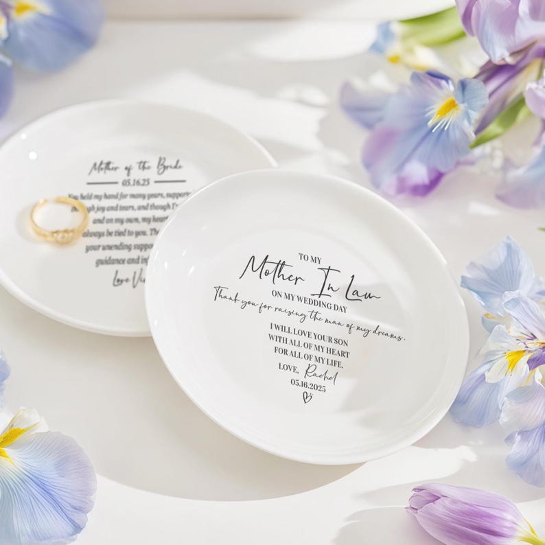Op de afbeelding: Twee witte keramische ringschalen met zwarte tekst. E&eacute;n leest "Mother of the Bride" met een datum. De andere zegt "Mother In Law" met een bericht en datum. Een gouden ring ligt op &eacute;&eacute;n schaal. Paarse bloemen omringen de schalen.