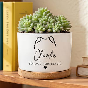 May include: White ceramic planter with a succulent plant. The planter features a black outline of a dog's face, the name "Charlie", the phrase "FOREVER IN OUR HEARTS", and a heart symbol. The planter sits on a wooden coaster.