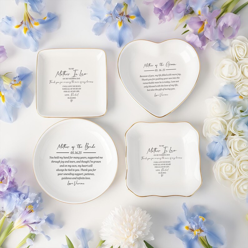 Op de afbeelding: Vier witte keramische schalen met gouden rand, elk met een andere vorm en inscriptie in zwart schrift. De tekst bevat zinnen als "Moeder van de bruid". Bloemsierkunst omringt de schalen.