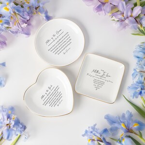 Op de afbeelding: Drie witte keramische schalen met gouden randen, in ronde, hart- en vierkante vorm. Elke schaal heeft zwarte tekst, waaronder "Mother of the Groom", "Mother of the Bride" en "Mother In Law". De schalen zijn omgeven door blauwe en paarse bloemen.