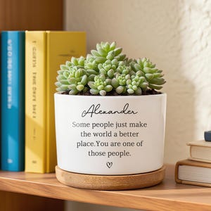 May include: A white ceramic planter with a succulent plant, sitting on a wooden coaster. The planter has the name "Alexander" and a message: "Some people just make the world a better place. You are one of those people."