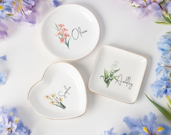May include: Three white ceramic dishes with gold rims, each featuring a floral design and a name in script: Olivia, Ashley, and Sophia. The dishes are round, square, and heart-shaped, respectively. Flowers and foliage surround the dishes.