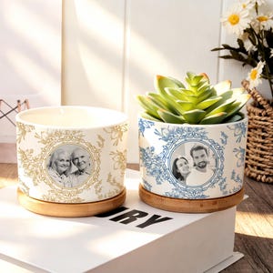 May include: Two cylindrical ceramic planters with bamboo saucers. The left planter features a gold floral pattern and a circular portrait of an older couple. The right planter has a blue floral pattern and a circular portrait of a younger couple.