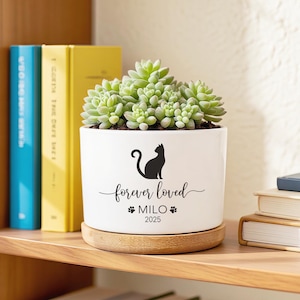 May include: White ceramic planter with a black cat silhouette, the words "forever loved", the name "Milo", and the year "2025". The planter holds a green succulent plant and sits on a wooden base. Books are in the background.