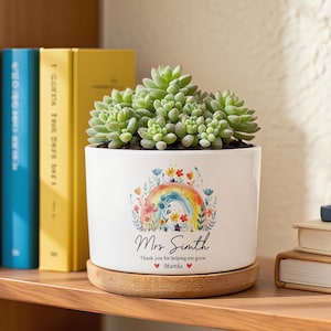 May include: A white ceramic planter with a rainbow and floral design, holding a green succulent plant. The planter has the text "Mrs. Simth" and "Thank you for helping me grow" with a heart symbol and the name "Martha". The planter sits on a wooden coaster.