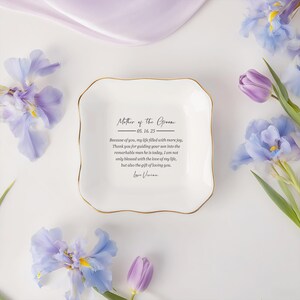 Op de afbeelding: Een wit keramisch schaaltje met een gouden rand, met de inscriptie "Mother of the Groom" en een oprecht bericht. Het schaaltje is omgeven door paarse en blauwe bloemen, wat een delicate en elegante esthetiek cre&euml;ert.
