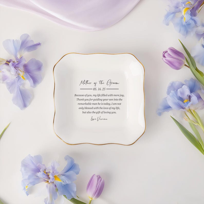 Op de afbeelding: Een wit keramisch schaaltje met een gouden rand, met de inscriptie "Mother of the Groom" en een oprecht bericht. Het schaaltje is omgeven door paarse en blauwe bloemen, wat een delicate en elegante esthetiek cre&euml;ert.