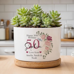 Peut inclure: Un pot de fleurs en céramique blanche avec une plante succulente. Le pot est décoré d'éléments floraux et du texte "Happy 50th Birthday Sophia" en noir et rose. Il repose sur une base en bois ronde.