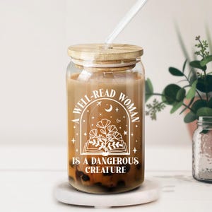 May include: A clear glass tumbler with a bamboo lid and a clear straw. The tumbler is filled with a beverage and features white text that reads "A WELL-READ WOMAN IS A DANGEROUS CREATURE" with floral and celestial designs.