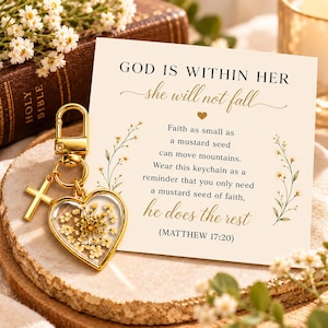 May include: A gold-toned keychain featuring a heart-shaped charm filled with pressed white flowers and a small cross charm. It is displayed on a cream-colored card with the text: GOD IS WITHIN HER she will not fall, Faith as small as a mustard seed can move mountains. Wear this keychain as a reminder that you only need a mustard seed of faith, he does the rest (MATTHEW 17:20).