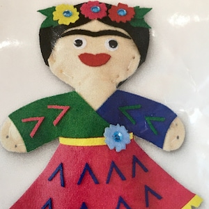 May include: Felt doll with a green and blue dress, a red skirt with blue triangles, and a flower crown with yellow and pink flowers. The doll has black hair and a painted face.