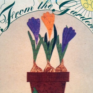 May include: A brown terracotta pot with three crocuses in bloom. The crocuses are purple, orange, and a darker purple. The pot is sitting on a beige background with the text "From the Garden" in green lettering.