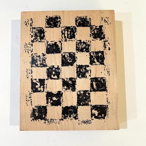 May include: A wooden stamp with a black and white checkerboard pattern. The stamp has a rectangular shape and is made of light-colored wood. The black squares have a textured appearance.