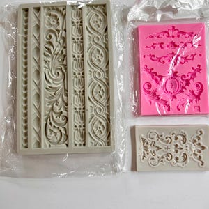 May include: Three silicone molds for crafting. One large rectangular gray mold features four distinct vertical decorative border patterns. A smaller rectangular pink mold displays ornate scrollwork designs. A small rectangular gray mold shows a central floral motif.