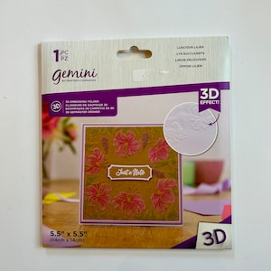 May include: A 3D embossing folder for crafting, featuring a design of pink lilies with the text "Just a Note". The folder is 5.5 inches by 5.5 inches.