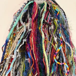May include: A bundle of assorted yarn strands in a wide variety of colors and textures, including fuzzy, metallic, braided, and smooth fibers. The strands are gathered at the top by a gray plastic hanger, creating a long, cascading fringe effect.